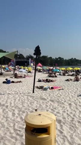 The video shows a crowded beach on a sunny day. White sand stretches across the foreground, with numerous beachgoers scattered across it, many lying on towels or under umbrellas. A tall, dark pole with a sign is prominently positioned in the middle ground, casting a shadow on the sand. In the background, a large white tent or canopy provides shade for a gathering of people. Further back, more tents and umbrellas are visible, along with a treeline bordering the beach. The sky is clear and blue. A man in swim trunks walks across the sand in the foreground. Several beach chairs, a cooler, and a trash can are also visible on the beach.