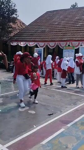 The video shows an outdoor scene likely at a school. The ground is paved and marked with white and colored lines. In the background, a building with a brown, tiled roof is visible, decorated with red and white banners. Many people are present, most wearing red shirts and white pants or skirts. Several individuals, particularly children, are wearing headscarves. In the foreground, a woman in a red jacket and white pants is walking with a young child. The child is wearing a pink skirt and carrying a bottle. Other children are moving around the scene, some with backpacks. The overall atmosphere appears to be an event or gathering.