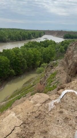 The video presents a wide shot of a river landscape in Meore Kesalo, Georgia, under an overcast sky. In the foreground, dry, cracked earth suggests a steep bank or cliff, with a discarded piece of white plastic visible on the ground. Below, a smaller, muddy-colored stream meanders through a lush green valley, lined with deciduous trees. This stream flows into a wider, light brown river, which is also flanked by dense green forests on both sides. A dirt path is visible on the left bank of the smaller stream. The scene captures the confluence of the two water bodies amidst natural vegetation and eroded terrain.