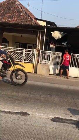 A dirt bike is ridden on a paved road in front of a house with a yellow gate. A person wearing a red shirt with the number 15 on the back is standing near the gate. The sun is shining and the sky is clear.