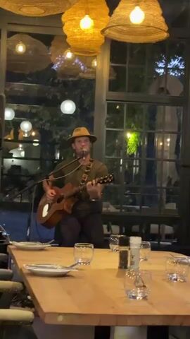 A man wearing a hat and holding an acoustic guitar performs in a restaurant. He is sitting in front of a microphone and facing the camera. He is wearing a khaki-colored shirt, dark pants, and a guitar strap. In front of him is a long wooden table with place settings for about 8 people. The place settings include plates, glasses, and silverware. There is a salt and pepper shaker. The restaurant is decorated with hanging woven light fixtures. Behind him is a window through which trees and string lights can be seen.