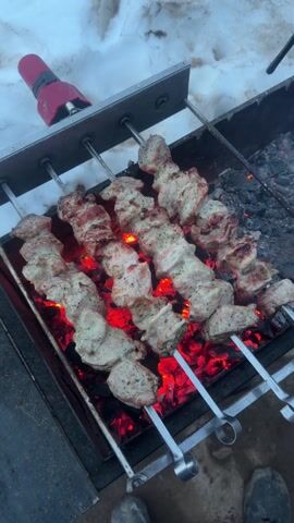 Four metal skewers loaded with pieces of meat are positioned over glowing-red coals in a metal grill. The skewers are held in place by a metal frame above the grill.  A small red electric lighter sits beside the frame.  To the upper left, a patch of snow can be seen.  A pair of shoes is barely visible at the bottom of the frame.