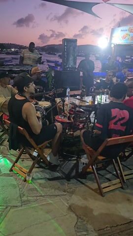 The video shows a group of musicians performing outdoors in front of a small crowd at dusk. The band, centered around a table filled with instruments and drinks, includes a vocalist playing guitar, a percussionist, and others. The audience sits on folding wooden chairs around the table, enjoying the music. A speaker stands near the table, amplifying the music. In the background, a harbor is visible, filled with small boats. A snack bar is also in the background with menus displayed. The setting suggests a relaxed, informal gathering enjoying live music by the water.