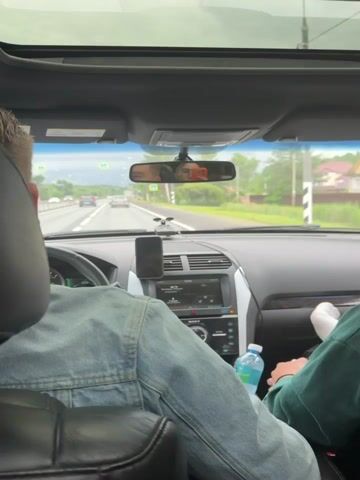 The video shows the interior of a car on a road trip. The camera is positioned behind the driver, looking forward through the windshield and at the rear-view mirror. In the driver's seat, a person wearing a denim jacket is visible from the back. Their left arm rests on the steering wheel, and their right hand is visible holding a bottle of water. In the passenger seat, another person's legs are visible, wearing a green sweatshirt and white socks.

In the rear-view mirror, two people are reflected. One, on the left, appears to be a young man with short blonde hair. The other, on the right, is a young woman wearing glasses and holding an orange smartphone. They seem to be taking a selfie or recording a video.

The scene outside the car is a two-lane road with other vehicles, including cars and a truck, traveling in the same direction. Trees and houses line the roadside, suggesting a rural or suburban setting. The sky is overcast, and the road appears wet, indicating recent rain or damp conditions.

The time displayed on the car's infotainment system is 13:01. The infotainment screen shows navigation information or media playback. A black smartphone is mounted on the dashboard in front of the infotainment system. A small black device, possibly a dashcam or radar detector, is mounted on the windshield above the infotainment system.

The overall mood seems relaxed and casual, typical of a car journey. The people are engaged in their own activities, with the woman in the mirror capturing a moment. The location is indicated as Mshaga, Russia.