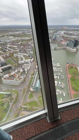 The video shows a view from a high-rise building overlooking a city. Through the angled windows, one can see a cityscape including buildings, roads, a river, a marina with boats, and some green spaces. The sky is overcast. The interior of the building is visible, with a dark colored floor and the supporting pillars of the building. There is a red light on one of the pillars.