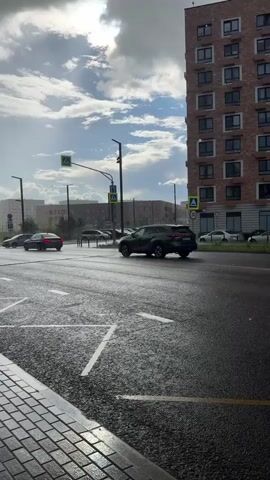 A rain shower is occurring in an urban environment. The camera is positioned low, looking across a wet, multi-lane road. The road surface has white painted markings, including lane dividers and a crosswalk. Several cars are driving on the road, appearing wet and reflecting light. One dark-colored SUV is in the foreground, moving away from the camera. In the background, more cars and buildings are visible. The sky is cloudy, with patches of sunlight breaking through, suggesting a break in the rain or a transition to clearer weather. The buildings are modern, tall, and appear to be residential or mixed-use. A street light with traffic signals is visible. The overall atmosphere is one of a city experiencing a brief, light rain shower.
