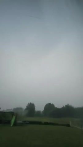 A short video clip shows a grey, overcast sky over a green lawn in Kyviškės, Lithuania. A children's slide and a trampoline are visible in the foreground on the left side of the frame. In the background, a line of trees forms a dark silhouette against the hazy sky. The weather appears to be rainy, with water droplets visible on what is presumably a windowpane through which the video is filmed. The overall mood is somber and quiet, typical of a stormy or foggy day. The time of day is not explicitly indicated, but the dim lighting suggests it is either early morning or late evening, or simply a very dark, cloudy day. There is no visible text in the clip.