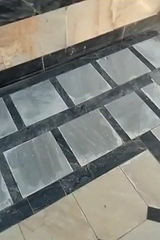 The video pans across a floor tiled with light and dark marble. The tiling alternates between square and rectangular patterns. The wall above the floor is also tiled with marble, featuring shades of beige and black. To the right of the frame, a metal gate with a diagonal pattern is visible. The gate appears to be closed, with a latch mechanism visible. Sunlight streams in from the right, illuminating the scene. A small metal bucket is partially visible in the lower right corner of the frame.