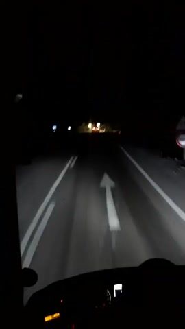The video shows a first-person perspective of driving a vehicle at night on a road. The headlights illuminate the road ahead, revealing white lines marking the lanes. Distant lights suggest other vehicles or structures. The dashboard of the vehicle is visible in the lower part of the frame, with some illuminated indicators. The surrounding environment is dark, indicating it is nighttime. The road appears to be paved and has visible tire tracks from previous vehicles. There are no visible people in the frame.