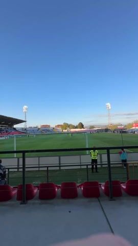 The video shows a sparsely populated stadium during a soccer game. The camera pans along a row of empty red seats, then up to a row of occupied seats, where two men are sitting. The stadium has large floodlights and a covered roof over the seating area. A few people can be seen on the field below. The sky is clear and blue.