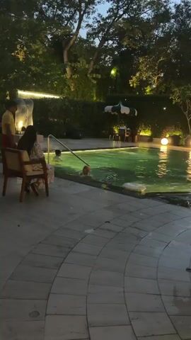 The video shows an outdoor swimming pool area at dusk. A woman is sitting on a chair on the left side of the frame, facing away from the camera. Two children are in the pool. One child, wearing a yellow shirt, is standing near a metal railing. Another child, wearing a pink swimsuit and an orange swim cap, is partially submerged in the water, holding something to their face. The pool water is illuminated with green and yellow lights. There are trees and bushes in the background, and a table with chairs and an umbrella is visible in the upper right corner. The ground around the pool is paved.