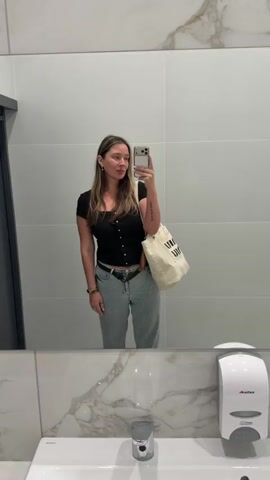 A woman with light brown hair, wearing a black top and light blue jeans, stands in front of a mirror and takes a selfie. She has a light-colored bag hanging from her left shoulder. She makes a few poses, smiles, and waves at the camera. The background is a white tiled wall with a mirror and a sink in the lower part of the frame.