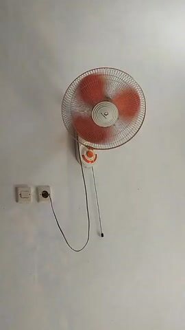 A wall-mounted oscillating fan with orange blades is shown from the front. The fan is plugged into a wall socket via a black cord. The fan is positioned on a white wall. To the left of the fan are two electrical outlets. The fan is operating, causing the blades to appear blurred. The video displays the fan from approximately the same angle throughout its duration.