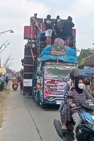 The video shows a flatbed truck adorned with a large sound system, including multiple speakers stacked on top of each other. Several people, some appearing to be performers or crew, stand on the elevated platforms of the truck, interacting with each other and the equipment. The truck is decorated with colorful banners and graphics. It is traveling down a paved road, which appears to be in a rural or semi-rural area, with buildings and trees visible on the sides. There are other vehicles and people present on the roadside, suggesting a public event or procession. The sky is overcast, and the overall atmosphere seems festive.