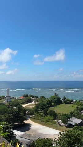 The camera pans left revealing a clear blue ocean, with a sandy bottom near the coast, under a blue sky with scattered white clouds. A sandy dirt road leads down to the beach area, with green trees and vegetation surrounding the road. Several small buildings and houses are visible among the trees. A larger parking lot with a lifeguard-style tower is also visible closer to the beach.