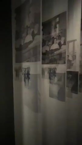 A video displays a wall covered with several black and white photographs arranged vertically on a white surface. The photographs depict groups of people, some appearing to be in formal settings or attending events. The images are somewhat faded and slightly blurry, with the lighting creating shadows and highlights across the display. The overall impression is of a collection of historical or personal memories.