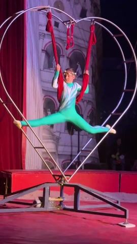 A young female acrobat is performing in a circus show. She is wearing a teal and white leotard. The acrobat is suspended in a large, heart-shaped metal apparatus with red fabric hanging from the top. The apparatus has a red base. The backdrop features a painting of a tower. The floor is red. The acrobat begins in a split position and then rotates to be upside down. A red curtain is visible in the background. Members of the audience can be seen in the dark foreground.
