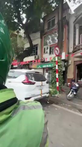 The video shows a person wearing a green helmet and a green jacket on a motorcycle riding through a city street. Buildings line the street, some decorated with red flags and banners. A white minivan passes to the right of the motorcycle. A Viettel Post office is visible.