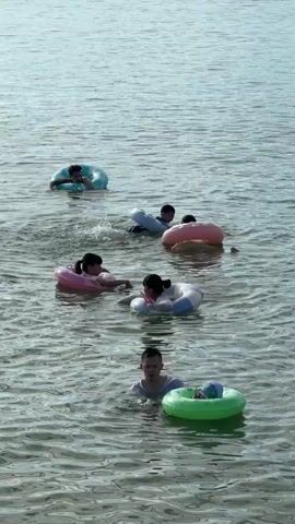 The video shows a group of people swimming in a body of water, appearing to be a lake or the sea. There are at least five individuals visible, all wearing swimming attire and using inflatable rings for buoyancy. The water is calm and appears to be shallow enough for people to stand in. The people are dispersed across the frame, with some closer to the foreground and others further in the distance. The lighting suggests it is daytime. The overall atmosphere is one of leisure and recreation.