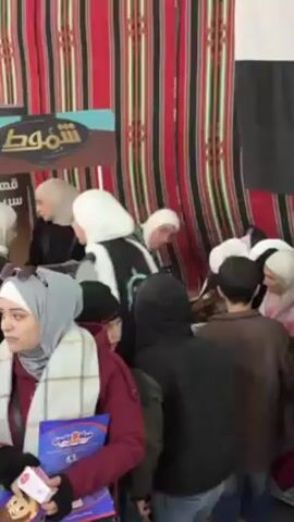 A group of people, including children, are gathered at a booth. Many of the women are wearing hijabs. The backdrop includes a flag with a red star and Arabic writing. A man in a black hoodie interacts with the children. There are signs with Arabic text visible. The environment appears to be a fair or community event.