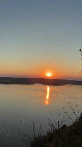 The video shows a scenic sunset over a body of water. The sun is a bright orange orb low on the horizon, casting a golden reflection on the water's surface. The sky transitions from a pale blue at the top to a warm orange and pink near the horizon. The water has gentle ripples, and a dark, silhouetted shoreline is visible in the distance. In the foreground, the edges of dry, leafless branches and some green foliage are visible, suggesting a high vantage point overlooking the landscape. The overall impression is one of tranquility and natural beauty as day transitions into evening.