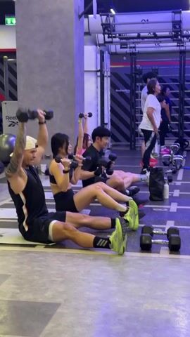The video shows a group of people exercising in a gym. The subjects are four individuals in the foreground, all seated on the floor with dumbbells. A man in a white hat, black tank top, and black shorts is on the left, performing a seated dumbbell press. To his right, a woman in a black sports bra and black shorts also holds dumbbells above her head. Next to her, a man in a black t-shirt and black shorts is doing the same exercise. Further to the right, a woman in a black sports bra and black shorts is also engaged in the exercise. In the background, several other people are visible, some standing and others exercising. The setting appears to be a modern gym with gray walls, industrial-style metal structures, and purple lighting. There are gym equipment like weights, yoga balls, and resistance bands. The floor has markings, suggesting it's a designated workout area. The activity is clearly a fitness class or group training session focused on strength training with dumbbells. The mood appears focused and energetic. The time of day is not explicitly indicated, but the lighting suggests it could be daytime or evening in an indoor environment. The location is identified as Nong Pa Khrang, Thailand. Visible text includes "Reebok" on a yoga ball and on a wall, and some Thai text on the wall in the background.