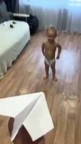 Here is a description of the video:
A baby in a diaper stands on a wood floor in a room, looking at the camera. A hand holding a white paper airplane throws it toward the baby. The baby laughs and walks away as the plane lands on the floor. The room has a bed, a black chair, a dresser, and a window with sheer white curtains.
