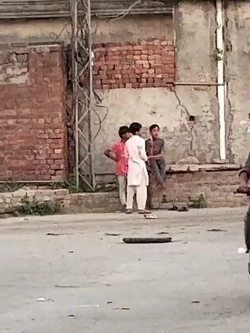 The video captures a candid street scene in Dera Chahal, Pakistan. In the background, three young boys are gathered near a dilapidated brick and concrete wall. One boy, wearing a red shirt, stands with his hands on his hips. Another, dressed in a white kurta, stands nearby, while a third boy in a darker shirt is seated on a low ledge against the wall. They appear to be conversing amongst themselves.

The foreground and midground show a paved street. A man on a motorcycle rides quickly from left to right, appearing blurred due to motion. To the far right, another man, partially visible, walks with what looks like a bicycle or motorcycle. In the immediate foreground, a boy in a plaid shirt stands with his back to the camera. On the ground, near the center, there is a flat, dark, circular object, possibly a discarded tire or part of one. The scene suggests a typical daytime setting in an urban or semi-urban environment.