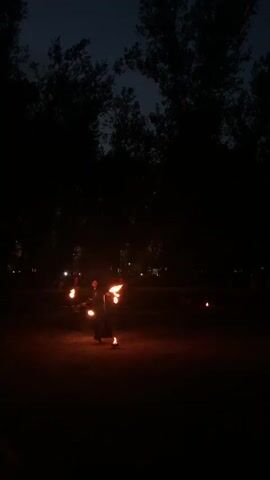 A person is performing a fire dance in a dark, outdoor area at night. They are holding multiple flaming props, likely poi, and are in motion. Several other small fires are visible on the ground in the background, and the silhouettes of trees are against the dark sky.