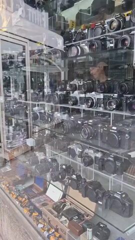 The video shows the exterior of a camera store. The store has large glass windows displaying a vast collection of cameras on shelves. The cameras vary in size and style, and some appear to be vintage. The store's signage is partially visible, with the words "Lab" and "-24 hour" discernible. The pavement in front of the store is visible, along with a concrete planter box. The overall impression is of a well-stocked store specializing in photographic equipment.
