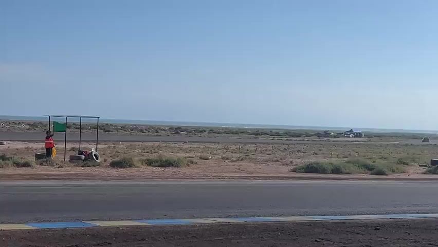 This media file captures a race taking place on a dry, dusty racetrack in Arica, Chile. The scene is set under a clear blue sky with a few wispy clouds, suggesting a warm, sunny day. In the background, the vast expanse of the Pacific Ocean is visible, with a ship on the horizon adding to the coastal context. The track itself is asphalt with yellow and blue painted markings. Surrounding the track is arid, desert-like terrain with sparse vegetation and a scattering of tires, typical of a racing venue in a dry climate.

The main activity is a car race, featuring several modified vehicles. A prominent red car, numbered "14" and with "FUMEY CUSTOM" text on its side, is a focal point, particularly in the early frames as it navigates a corner. Later, a yellow and black car with the number "114" and various decals, including what appears to be a tiger graphic, is shown in action, emitting a puff of smoke as it drifts around a turn. Other cars, including a white hatchback, are also visible on the track and in the pit area.

A person in an orange vest and a dark hat stands near a temporary structure on the side of the track, holding a green flag, likely a race official or marshal. Another person in an orange vest is visible further down the track. The atmosphere appears to be one of competitive sporting activity. The time of day is clearly daytime, judging by the bright sunlight and the long shadows cast. The overall impression is a low-budget, spirited racing event in a unique coastal desert environment.