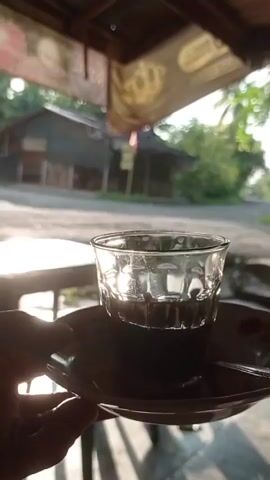 A close-up shot shows a hand holding a saucer with a glass of dark liquid, likely coffee, on it. The sun is shining brightly, creating a lens flare effect on the left side of the image. The background is blurred and out of focus, showing some buildings and trees. The scene appears to be outdoors, possibly at a roadside stall or cafe.