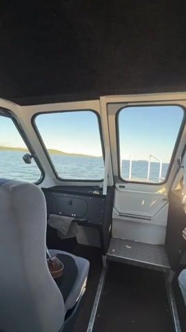 This media file provides a first-person perspective from inside a boat, likely a cabin cruiser, as it travels across calm waters. The view shifts between looking forward through the front windows and to the sides.

In the forward view, a person wearing a light gray hoodie and light-colored pants is at the helm, steering the boat. Sunlight streams in from the right, illuminating the interior. Through the windows, a vast expanse of water is visible, meeting a tree-lined shoreline under a clear, bright blue sky. The time of day appears to be late afternoon or early evening, given the warm, angled sunlight. The overall mood conveyed is one of peaceful travel and enjoyment of a scenic natural setting.

The interior of the boat is functional, with dark carpeting on the ceiling and floor, and white walls and window frames. A black dashboard with some controls is visible on the left, with a folded towel placed on it. A grey and black seat is in the foreground on the left. A metal step leads to the exit at the rear.

The side views through the windows reveal the same tranquil water and forested coastline. The gentle ripples on the water's surface suggest a calm day with minimal wind. There are no visible signs or text within the media. The setting is clearly a body of water surrounded by natural, undeveloped land, consistent with a location like Tarulinna, Russia, which is known for its natural beauty.