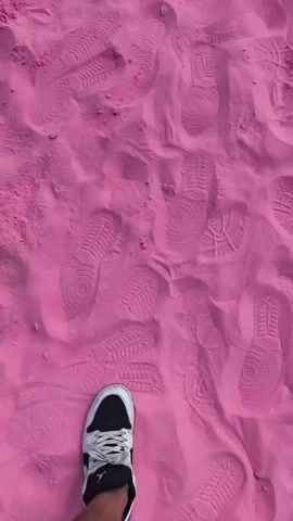 A person's feet are shown walking on bright pink sand. The sand is disturbed by numerous footprints, indicating previous activity. The person is wearing white sneakers with black accents and black shorts. The camera angle is overhead, focusing on the sand and the person's legs and feet.