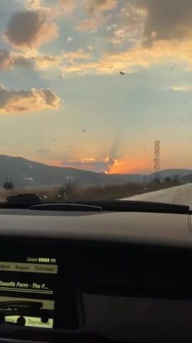 The video is filmed from inside a car looking out through the windshield. The car appears to be driving on a paved road through a rural landscape during sunset. The sky is filled with dramatic clouds illuminated by the setting sun, casting orange and yellow hues. Sunlight beams through the clouds, creating a crepuscular effect. Mountains are visible in the background, and a tall communication tower stands on the right side of the road. The dashboard of the car is visible in the foreground, showing a car infotainment system with Russian text displayed. There are small specks on the windshield, possibly bugs or dust, which are illuminated by the sunlight. The overall impression is one of a scenic drive at dusk.