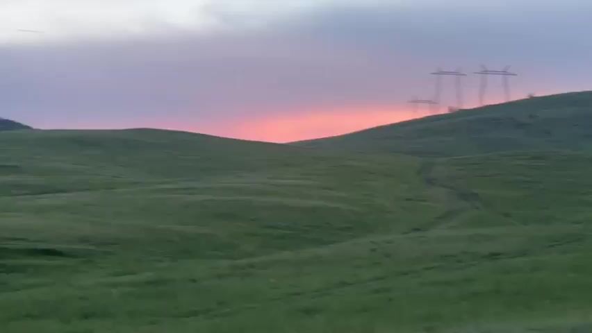 The video shows a panoramic view of a rolling green hills landscape at dusk. The sky is painted with soft hues of pink, purple, and grey, indicating the setting sun. Several tall electrical transmission towers with wires are visible on the horizon, silhouetted against the colorful sky. A faint dirt track winds its way up one of the hills. A few scattered trees dot the landscape, some grouped together and others standing alone. The grass on the hills is short and appears to be uniformly green, suggesting it is a pasture or meadow. The camera is panning slowly from left to right, offering a broad view of the serene, open scenery. There are no people visible in the video.