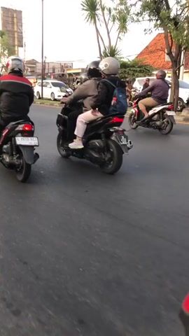 The video shows a street scene with several motorcycles and cars. In the foreground, a motorcycle is being ridden, and the side mirror reflects other vehicles. In the middle ground, several motorcycles are in motion, with riders wearing helmets. One motorcycle has two people on it, one driving and one passenger. Another motorcycle has a single rider. In the background, there are buildings, trees, and a white SUV. The sky is overcast. The overall impression is of a busy street with ongoing traffic.