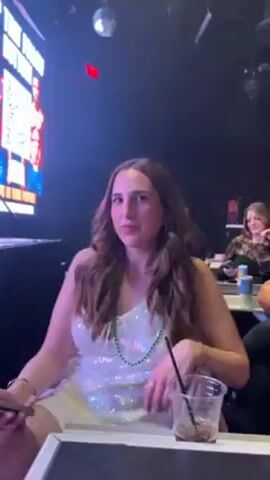A woman with brown hair wearing a shiny white dress sits at a table in a dim nightclub. She has a green necklace and is waving her hand.  A plastic cup with a straw sits on the table in front of her. A woman sits at a table behind her. A screen with a poster is visible on the left. A disco ball hangs from the ceiling.