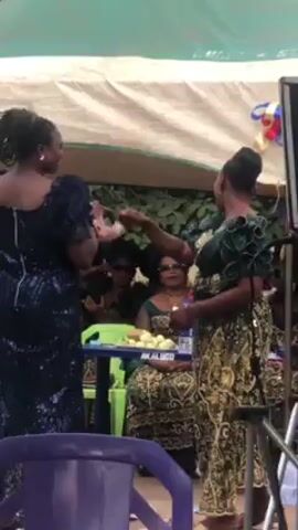 A woman in a dark blue, sequined dress is standing and speaking at what appears to be an outdoor event. She is holding a small, rectangular item in her hands, possibly a booklet or card. Behind her, several women are seated at a table. The table has a sign on it that reads “AKALUCO.” There are also water bottles and a plate of what looks like apples on the table. A partially visible motorcycle sits to the left of the table. The event appears to be taking place under a white tent or canopy, and there is some greenery visible in the background. Other people are standing and sitting around the area, observing the woman speaking.