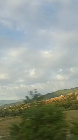 The video shows a landscape as viewed from a vehicle in motion. The sky is filled with clouds, with patches of blue visible. Below the sky are rolling hills covered in greenery, with some bare, dry patches on the slopes. In the foreground, there is a field and what appears to be a road or highway with a guardrail. A light pole is visible on the left side of the frame. The scene suggests a rural or semi-rural environment, possibly during the daytime with overcast or partly cloudy conditions.