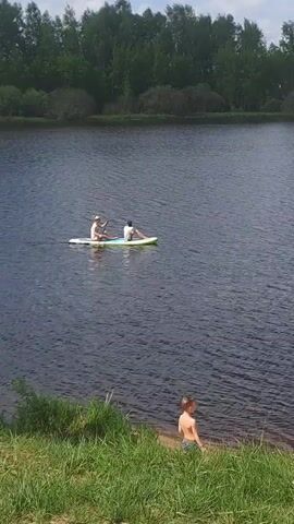 A video shows a woman and a young person on a stand-up paddleboard on a body of water. The woman is paddling the board while the young person sits facing the direction of travel. A young boy stands on the grassy shore. The water is rippled and dark in color, and dense trees are visible in the background. The sky is partly cloudy.