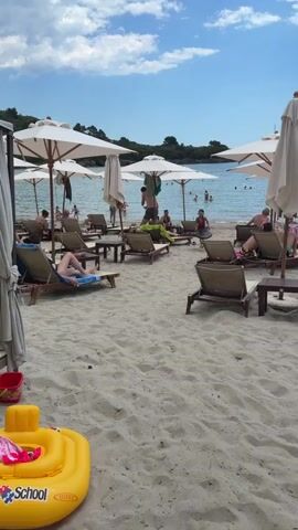 This media file depicts a sunny day at a beach in Milovići, Montenegro. The scene is filled with people enjoying a leisurely afternoon. Several beachgoers are relaxing on lounge chairs under large white umbrellas, while others are in the water, swimming or wading.

A young girl is the central focus of the activity, running across the sand with a boogie board. She appears to be excited and energetic. In the foreground, a bright yellow inflatable toy, possibly a ring float, is partially visible, suggesting a family-friendly environment. The sand is light-colored and appears well-maintained.

The water is calm and blue, with distant trees and land visible on the horizon. The sky is a clear blue with scattered white clouds, indicating good weather. The presence of umbrellas and lounge chairs suggests that this is a well-equipped beach, possibly with amenities for visitors. The overall atmosphere is one of relaxation and vacation enjoyment.

There is some visible text on the yellow inflatable toy, but it is partially obscured and difficult to read clearly. It appears to be part of a logo or brand name. The time of day is likely midday or early afternoon, given the bright sunlight and the activities taking place.