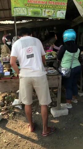 The video shows a street-side vendor stall. A man in a white t-shirt and tan shorts is standing at the stall, facing away from the camera. He appears to be looking at items on display. A woman wearing a light green shirt, dark pants, and a blue helmet is standing to the right of the man, also looking towards the stall. Behind the counter, another person, likely the vendor, is partially visible. The stall has various items for sale, including what appear to be packaged goods and possibly food items. The environment is outdoors, with dirt ground and some vegetation visible. The lighting suggests it is daytime. The overall scene depicts a casual transaction at a local market or roadside shop.