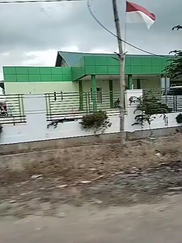 The camera is mounted on a vehicle moving along a road, capturing a view of a building. The building is a light green color with a white trim and a green roof. There is a sign that reads "Rumah Sakit Zahra" in white letters. A tall pole with an Indonesian flag is visible in front of the building. There are trees and foliage in front of the building. The sky is overcast, and the ground appears dry and dusty. As the vehicle approaches the building, the view switches to the entrance area with the letters "IGD 24 JAM" above it. A white vehicle is visible within the entrance area.