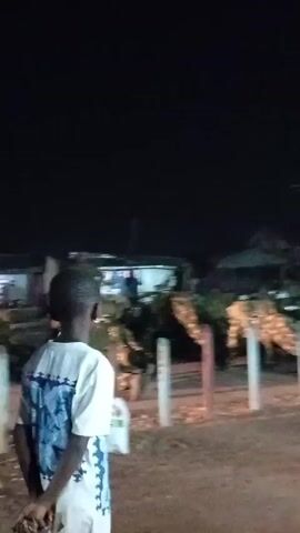 At night, a child stands with their back to the camera, observing a group of people in camouflage uniforms performing exercises near a small building. The people are performing push-ups near a row of short, white and blue posts. Debris and a grey bucket are in the foreground near the posts. A person is standing under the bright lights of the building. The environment appears to be outdoors in a semi-urban setting.