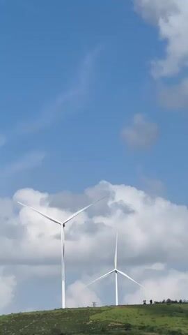 The video shows two wind turbines on a grassy hill under a blue sky with white clouds. The wind turbines are white and tall with three blades each. The blades of the turbines are spinning. The hill is covered in green grass and some trees or bushes are visible on the right side of the frame. The sky is mostly blue with some scattered clouds. The footage is taken from a distance, looking up at the turbines.