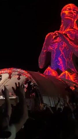 The video shows a dark outdoor scene at night, likely a concert or festival. A large, illuminated sculpture of a seated figure, possibly a woman, is prominently displayed in the background. The sculpture is projected with vibrant, colorful imagery, appearing to depict an underwater scene with aquatic plants and fish. In the foreground, a stage structure is visible, illuminated by spotlights. A DJ or performer is seen behind a console on the stage. A crowd of people are gathered in front of the stage, with many raising their hands in the air, suggesting they are enjoying the performance. The overall atmosphere is one of excitement and entertainment.