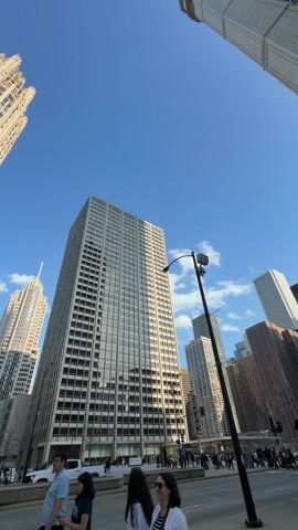 The video shows a street in Chicago. Several people walk on the sidewalk. The camera focuses on the tall skyscrapers on either side of the street, towering towards a clear, bright blue sky. The architectural styles of the buildings vary, with some appearing modern and others displaying more historical designs. A bird flies in the sky between buildings. Traffic moves along the street below. The overall scene captures the urban landscape of a bustling city center.