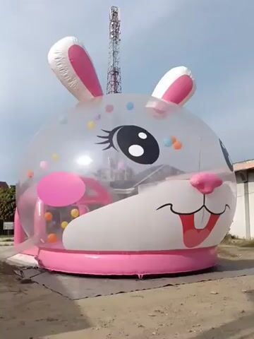 A clear, round inflatable bunny structure with pink accents and a cartoon face sits on a patch of dirt or sand. The bunny has large, black eyes with eyelashes, a pink nose, and an open mouth with a pink tongue and white teeth. Inside the clear bubble, there are small, colorful balls. Pink inner structures are also visible. The structure is set up outdoors, with a cell tower and some buildings in the background under a bright sky. The camera appears to be moving slightly, showing the bunny from different angles.