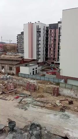 The video shows a construction site with several multi-story apartment buildings in the background. The buildings are modern in design with a combination of white, pink, and gray facades, some with balconies. Between the camera position and the buildings is a construction site full of debris, including wooden planks, pallets, and metal containers.  There is also a small, older industrial building with a rusty roof and a newly built small white building.  Beyond the construction site, there is a playground with a slide and other equipment. In the distance, there is a construction crane visible against a cloudy sky.